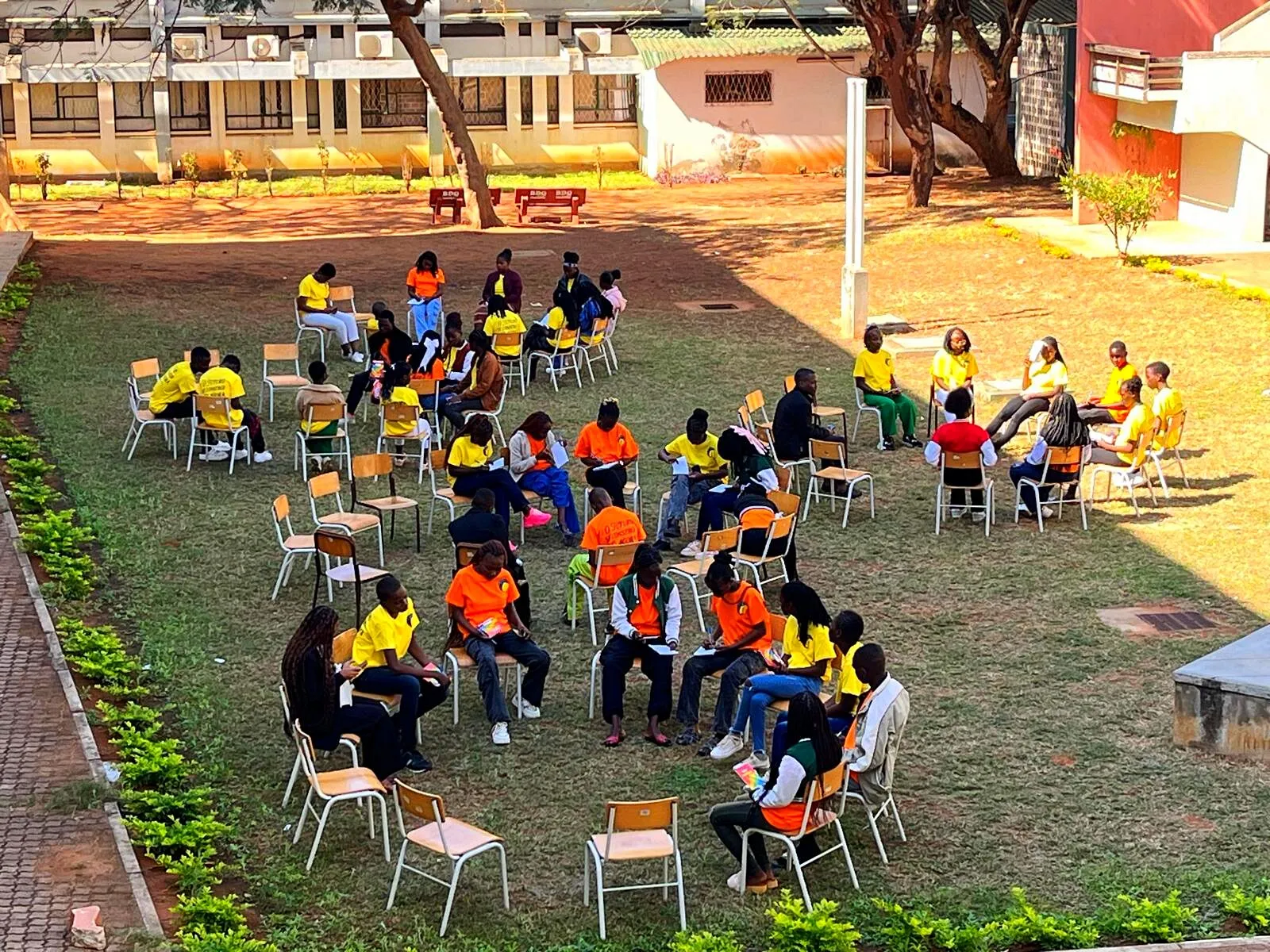 Vistos de cima, cerca de quarenta jovens de T-shirt amarela e laranja sentados em cadeiras em pequenos círculos num pátio relvado