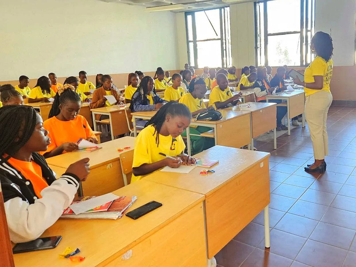 Duas facilitadoras de T-shirt amarela à frente de uma sala de aula cheia de alunos de amarelo e laranja, a escrever nas carteiras