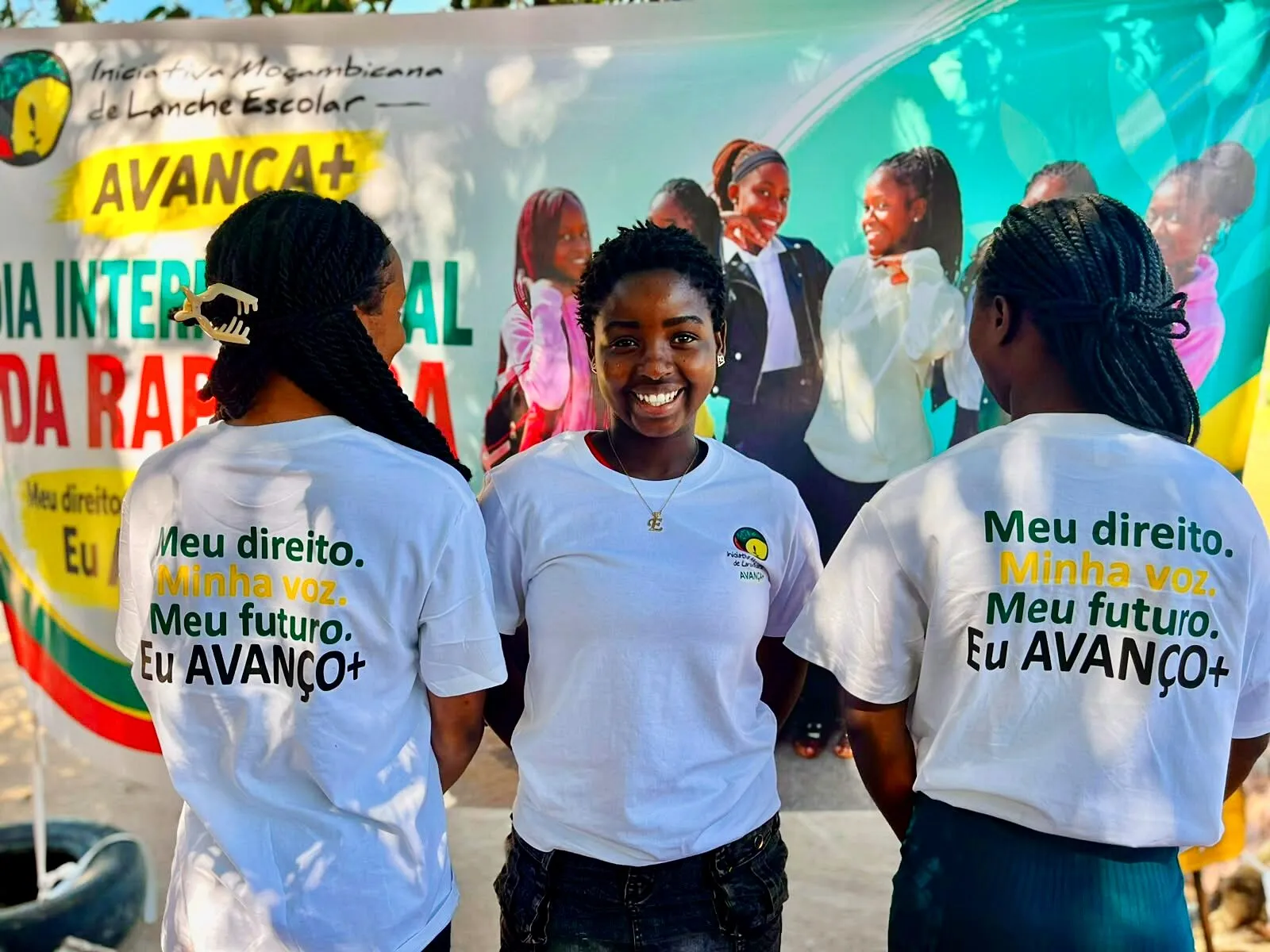 Três raparigas adolescentes de T-shirt branca do Avança+ diante de uma faixa do Dia Internacional da Rapariga; a do meio sorri para a câmara