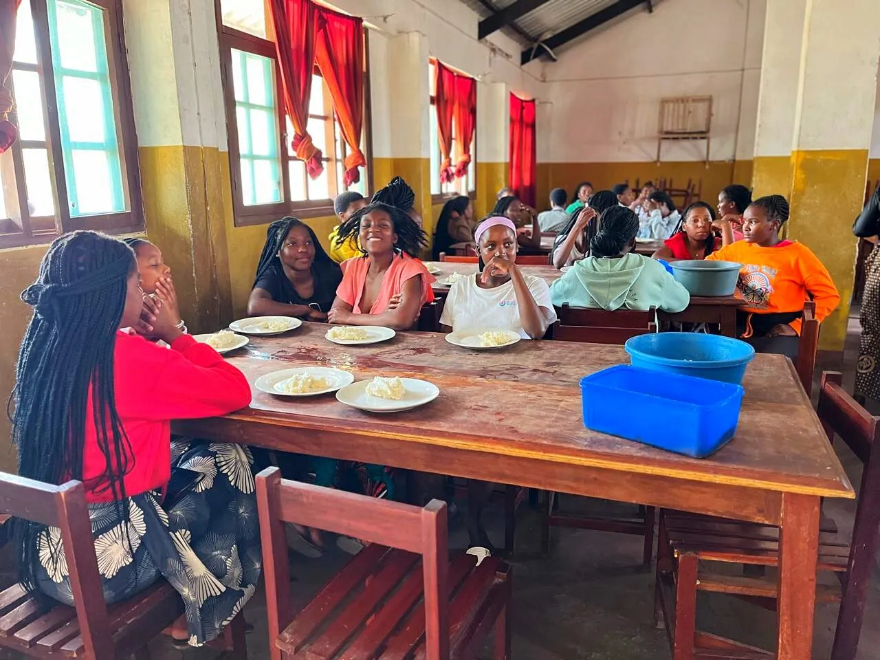 Raparigas adolescentes em mesas compridas de madeira num refeitório, a comer xima em pratos brancos e a rir