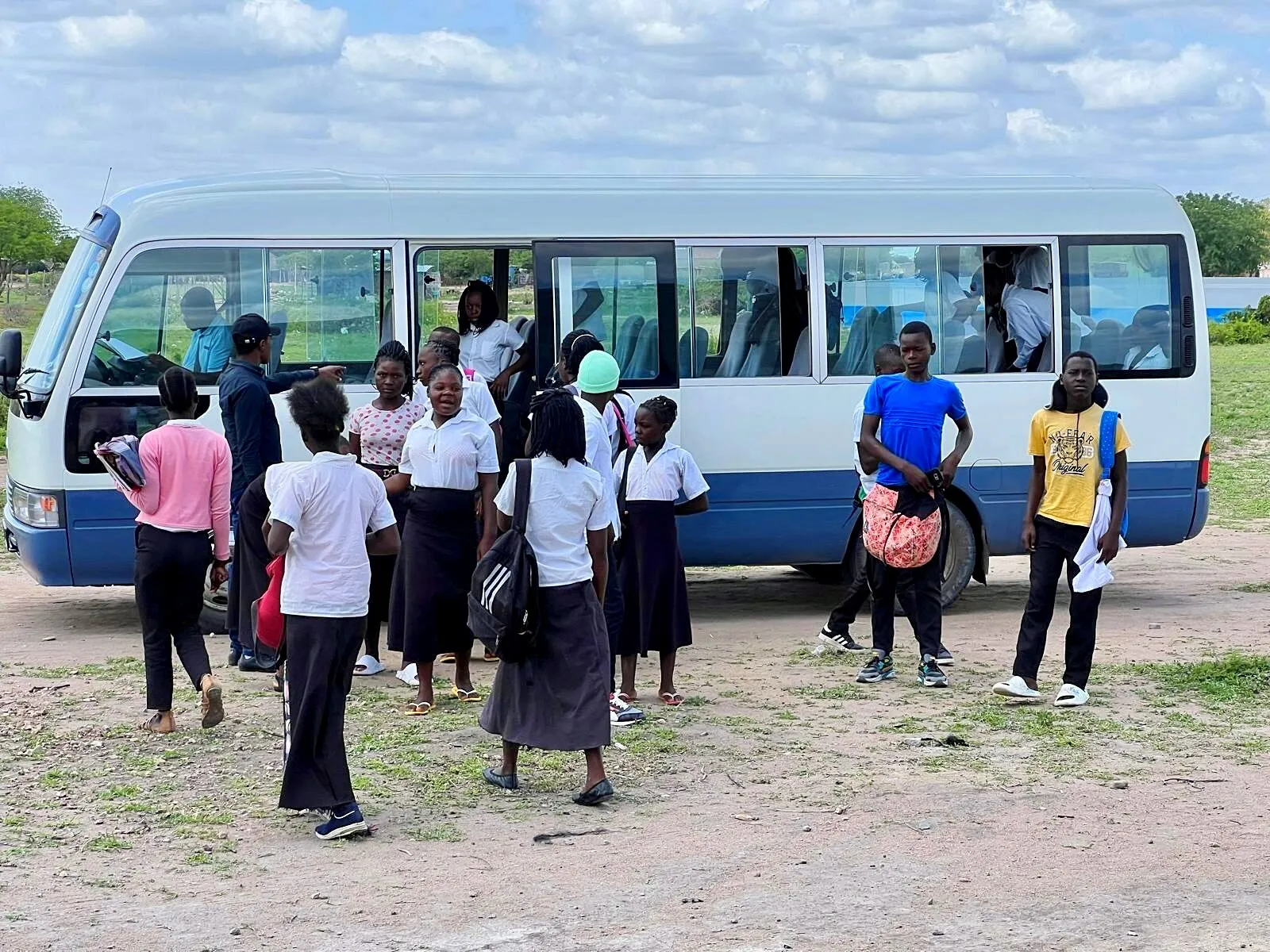 Cerca de quinze alunos do secundário de camisa branca, com mochilas, junto à porta lateral aberta de um minibus branco e azul
