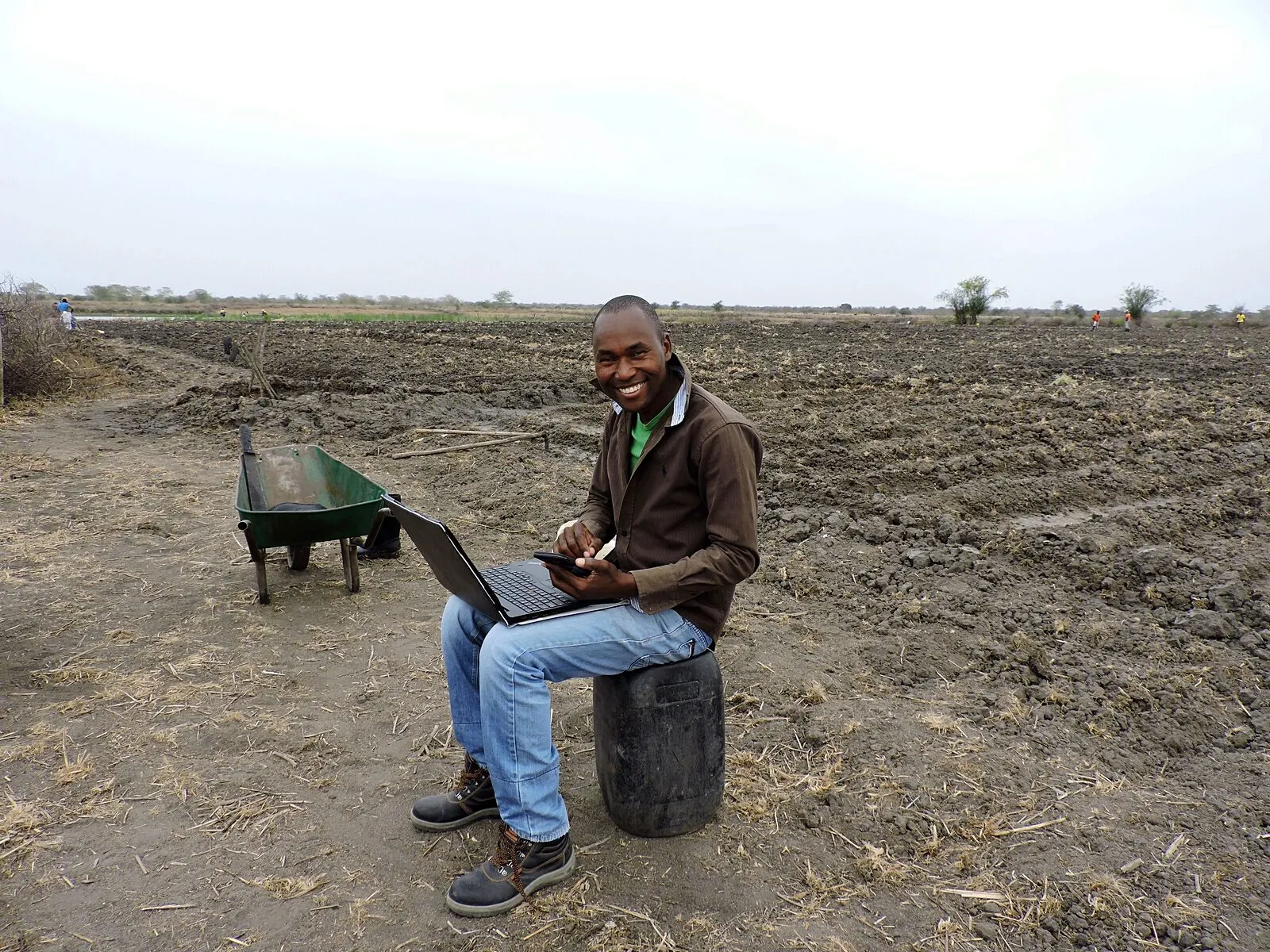 Roberto Mutisse sentado num bidão de plástico virado ao contrário, num campo agrícola lavrado e seco, sorridente, com um portátil sobre os joelhos e um carrinho de mão verde ao lado