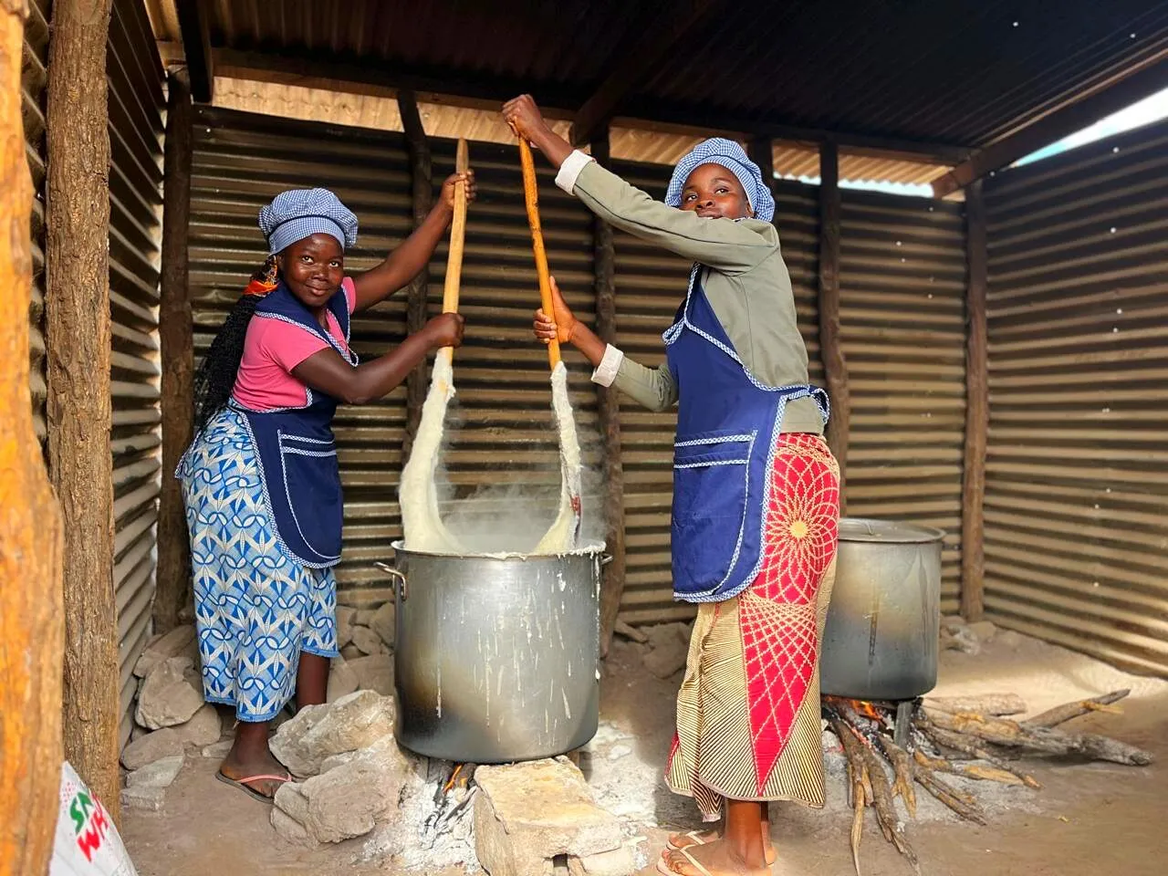 Duas cozinheiras de touca aos quadrados mexem panelas de xima com pás compridas numa cozinha escolar de chapa