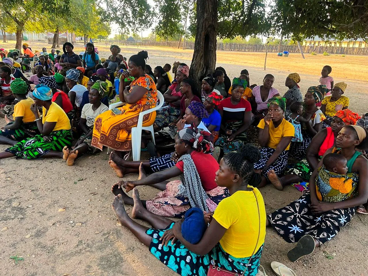 Cerca de sessenta mulheres, muitas com bebés e crianças pequenas, sentadas na areia e em cadeiras de plástico à sombra de uma grande árvore, numa reunião comunitária