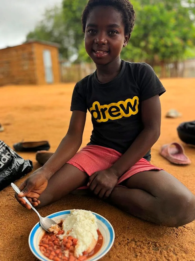 Uma menina sentada na areia vermelha com uma tigela de xima e feijão, a sorrir para a câmara