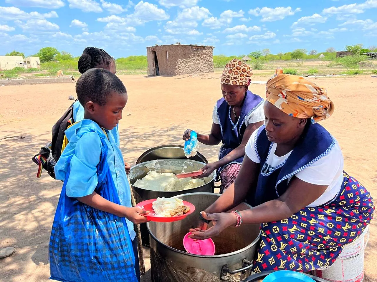 Duas cozinheiras servem xima de uma panela grande no prato de uma menina de uniforme escolar azul