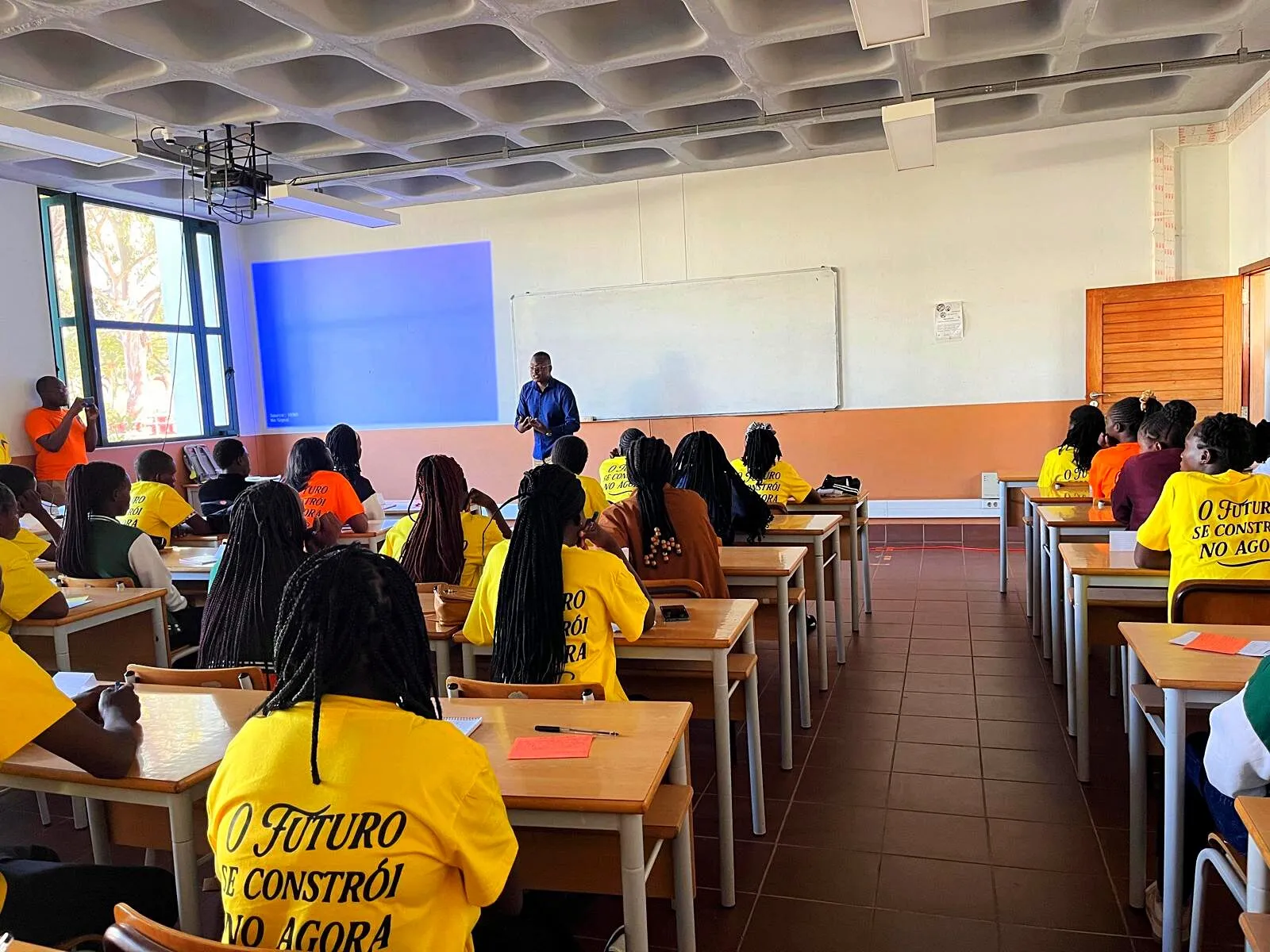 Do fundo de uma sala de aula universitária, filas de raparigas de T-shirt amarela e laranja com a frase "O futuro se constrói no agora" ouvem um homem que fala à frente