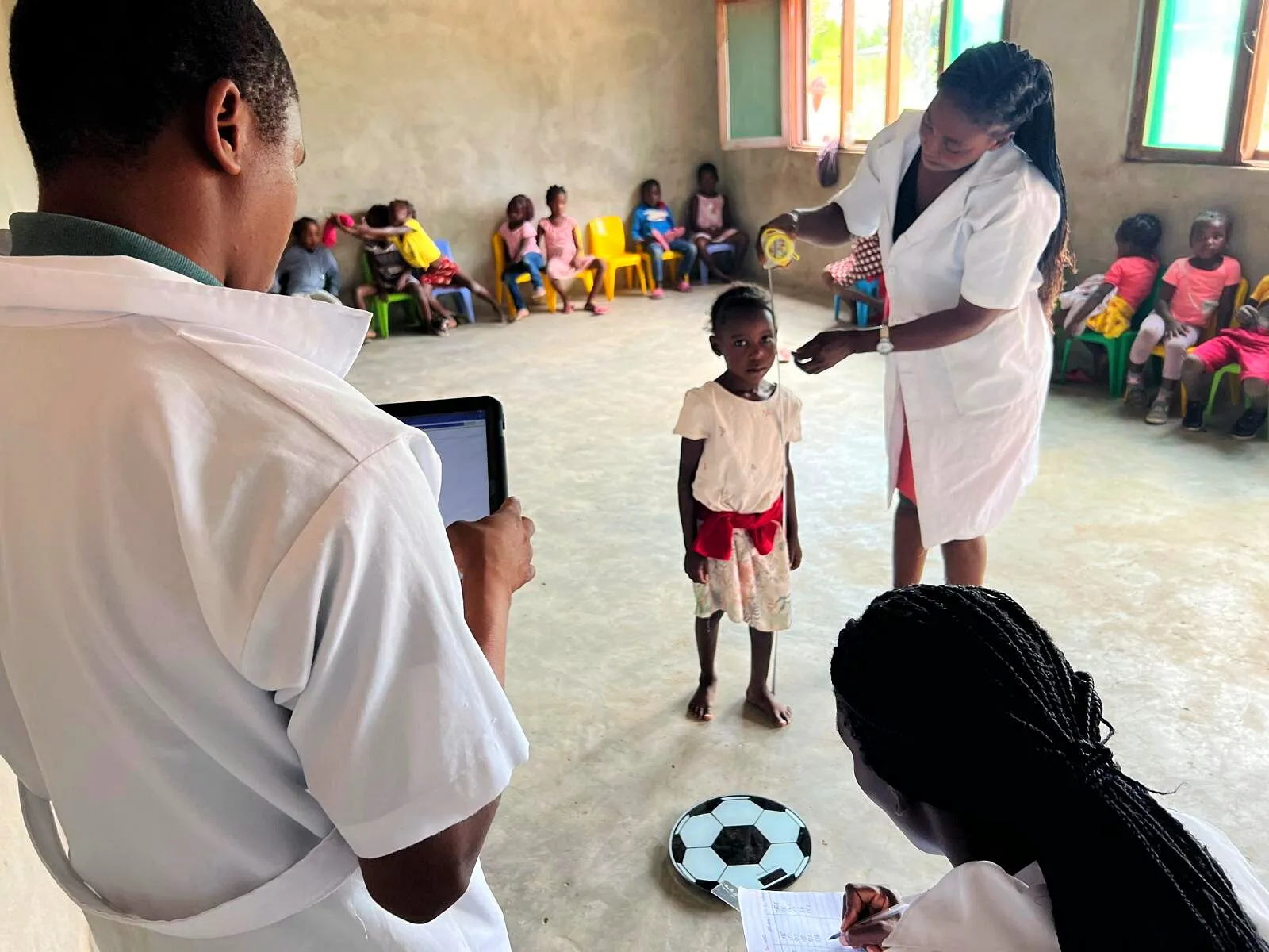 Numa sala de aula, uma mulher de bata branca mede a altura de uma menina pequena enquanto colegas registam os dados e outras crianças esperam