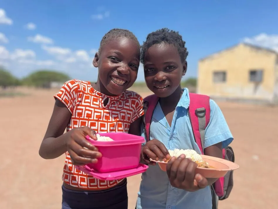 Duas meninas sorridentes num pátio escolar mostram o seu almoço de arroz e caril, uma numa caixa cor-de-rosa, outra num prato