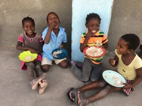 Alunos pequenos sentados encostados a uma parede a comer dos pratos