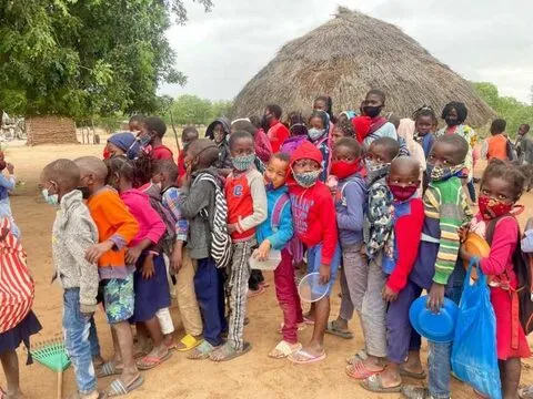 Uma longa fila de alunos com pratos ao lado de uma palhota