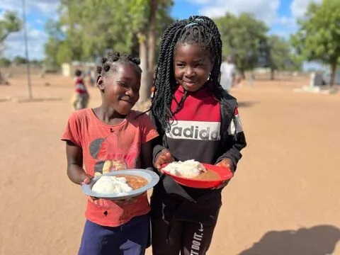 Duas meninas seguram pratos de arroz num pátio escolar de areia