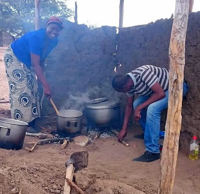 Uma cozinheira sorridente mexe uma panela sobre uma fogueira junto a um muro de barro, enquanto um homem atiça o lume sob uma segunda panela