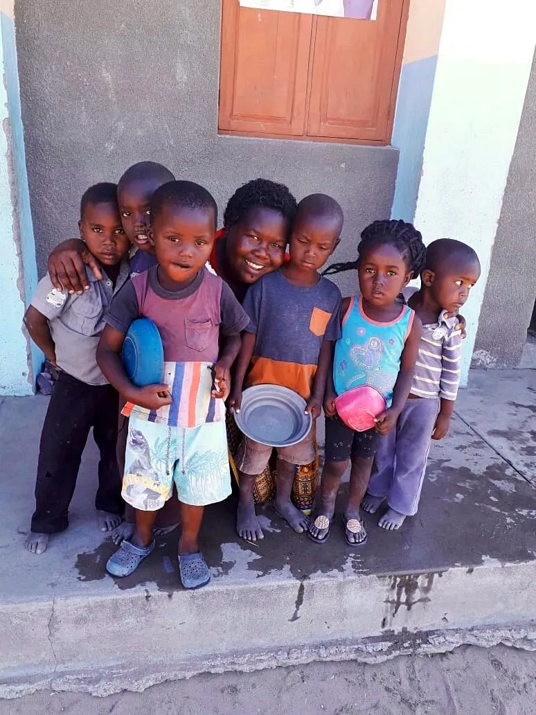 Talvina Ualane agachada e sorridente atrás de seis crianças pequenas com pratos de plástico, no degrau de um edifício escolar cinzento e azul