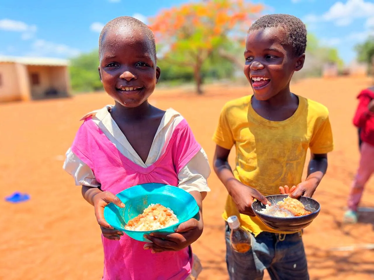 Uma menina sorridente e um rapaz a rir estendem tigelas de arroz e feijão sobre terra vermelha, com uma acácia-rubra em flor atrás