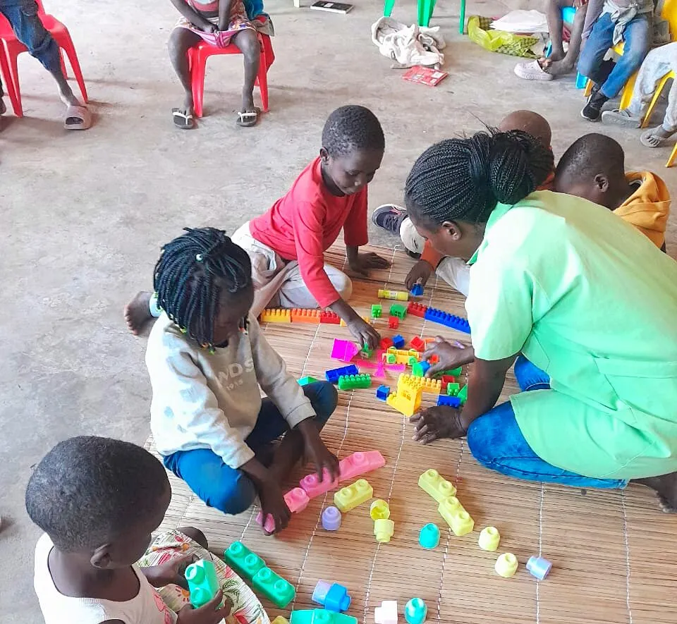 Uma educadora de bata verde-clara ajoelhada numa esteira com crianças pequenas a construir com blocos de plástico coloridos