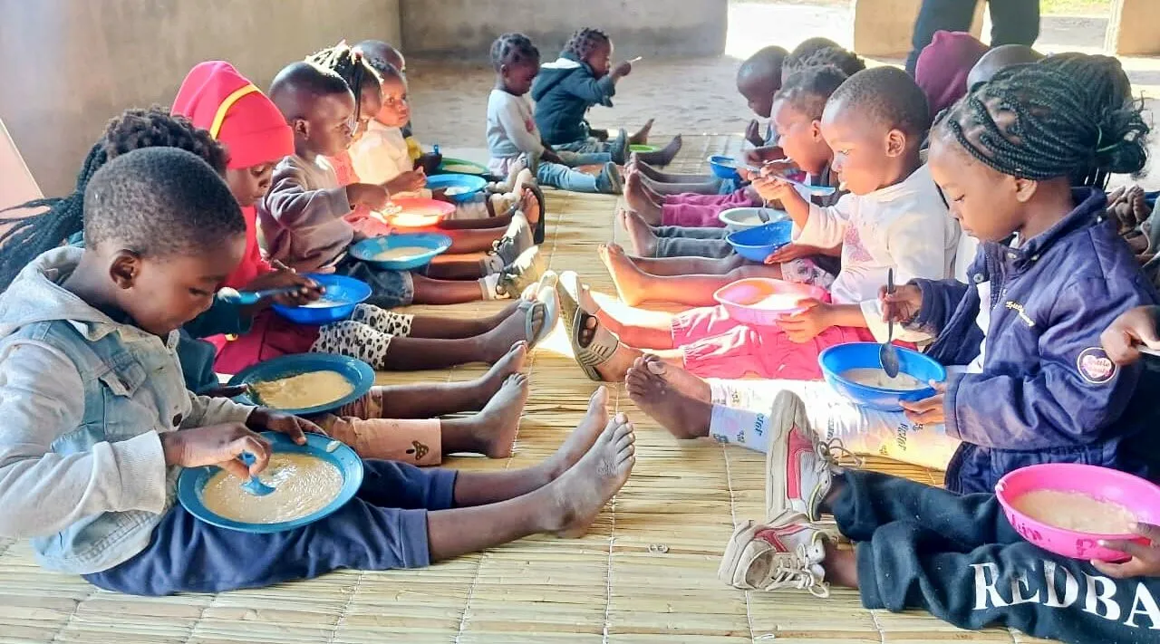 Duas filas de crianças de uma escolinha comunitária sentadas frente a frente numa longa esteira, a comer papa em tigelas azuis e cor-de-rosa debaixo de um alpendre de cimento