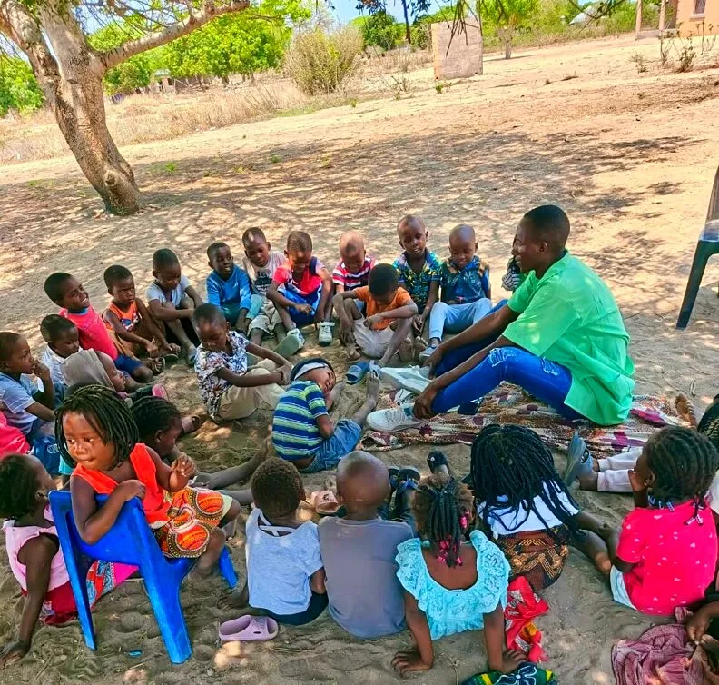 Um jovem de bata verde-clara sentado debaixo de uma árvore a ler para cerca de vinte e cinco crianças de uma escolinha comunitária sentadas em roda na areia