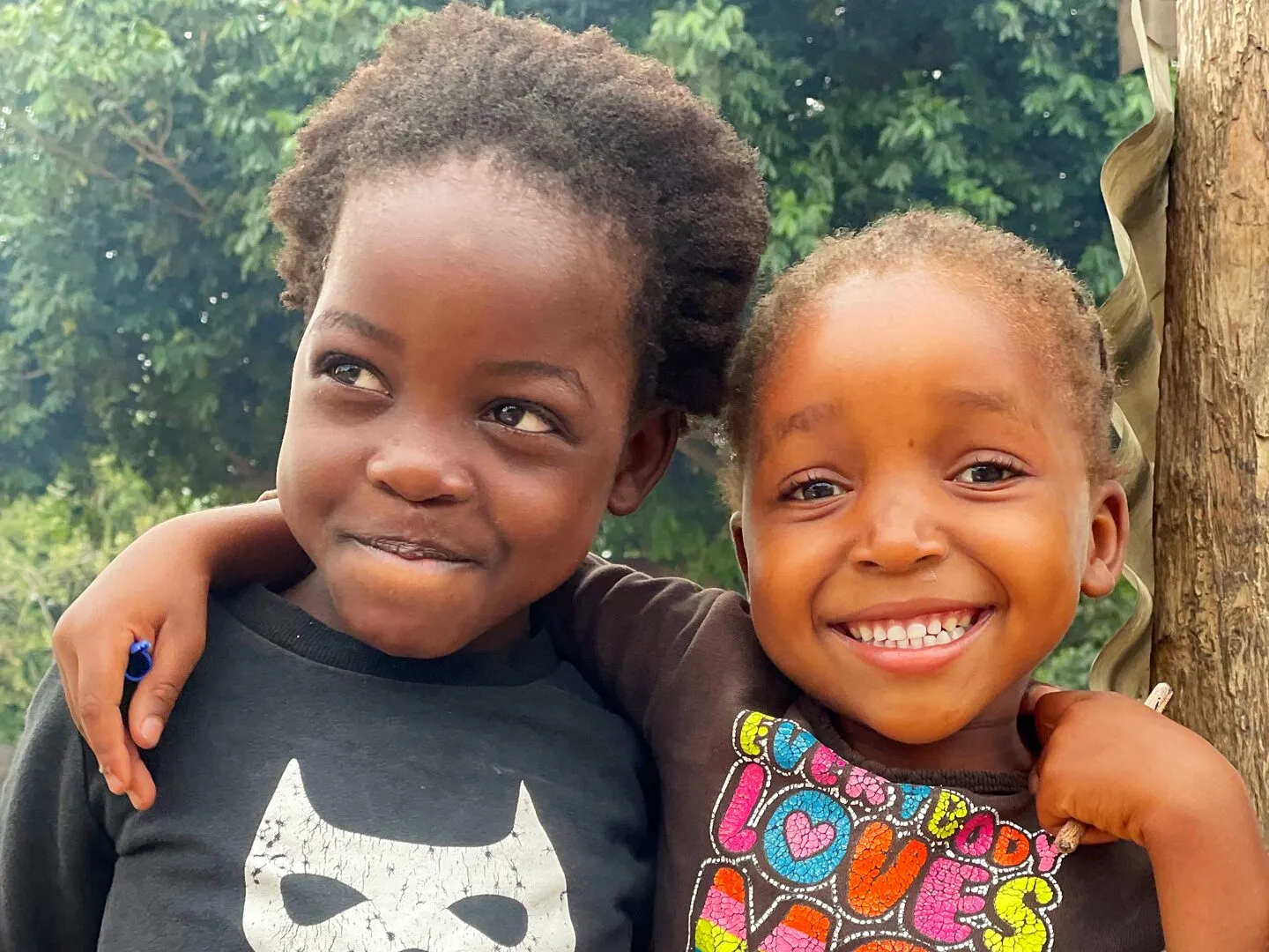 Duas meninas pequenas e sorridentes, de camisolas escuras, abraçadas diante de folhagem verde