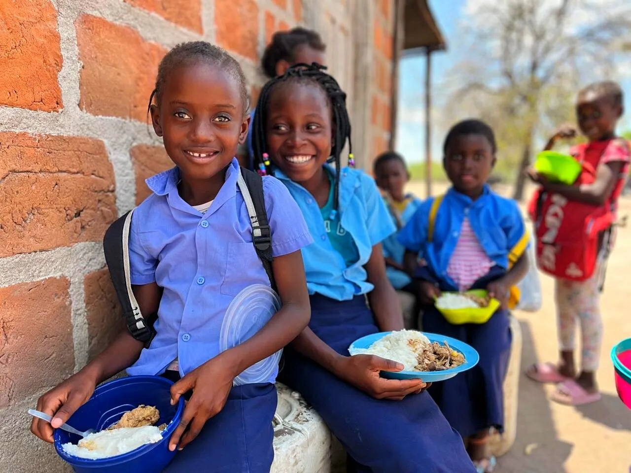 Duas alunas sorridentes de uniforme azul sentadas junto a uma parede de tijolo a almoçar xima com caril de uma tigela e de um prato