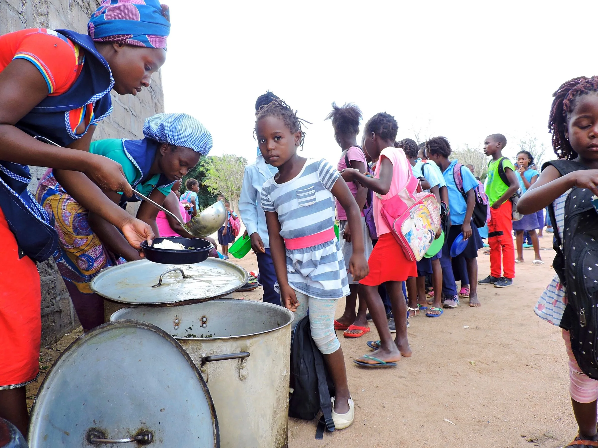 Duas cozinheiras de lenço axadrezado servem o almoço de uma panela grande para a tigela de uma criança, enquanto uma longa fila de alunos espera junto à parede da escola