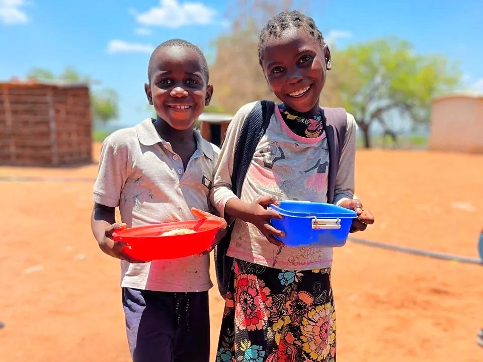 Um menino sorridente com uma tigela vermelha de comida e uma menina sorridente de mochila com uma marmita azul, lado a lado na areia vermelha