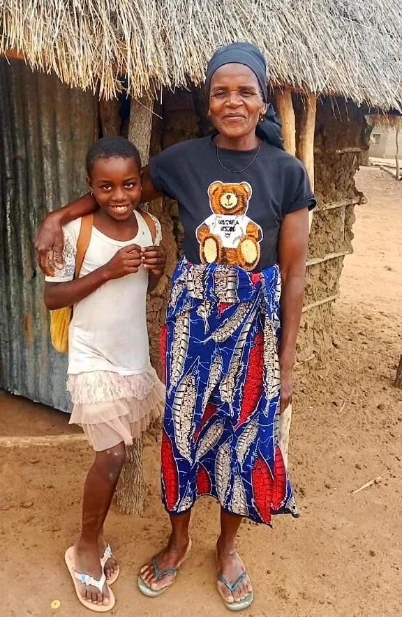 A Hortencia, uma menina de blusa branca, abraça uma senhora mais velha diante de uma casa de capim