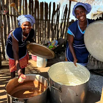 Duas cozinheiras de avental azul e lenço na cabeça riem-se junto a duas panelas grandes de comida