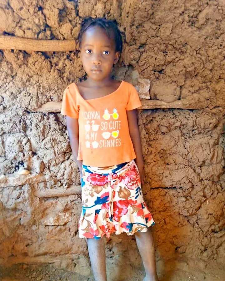 Uma menina de t-shirt cor de laranja e saia florida, de pé encostada a uma parede de barro, a olhar para a câmara