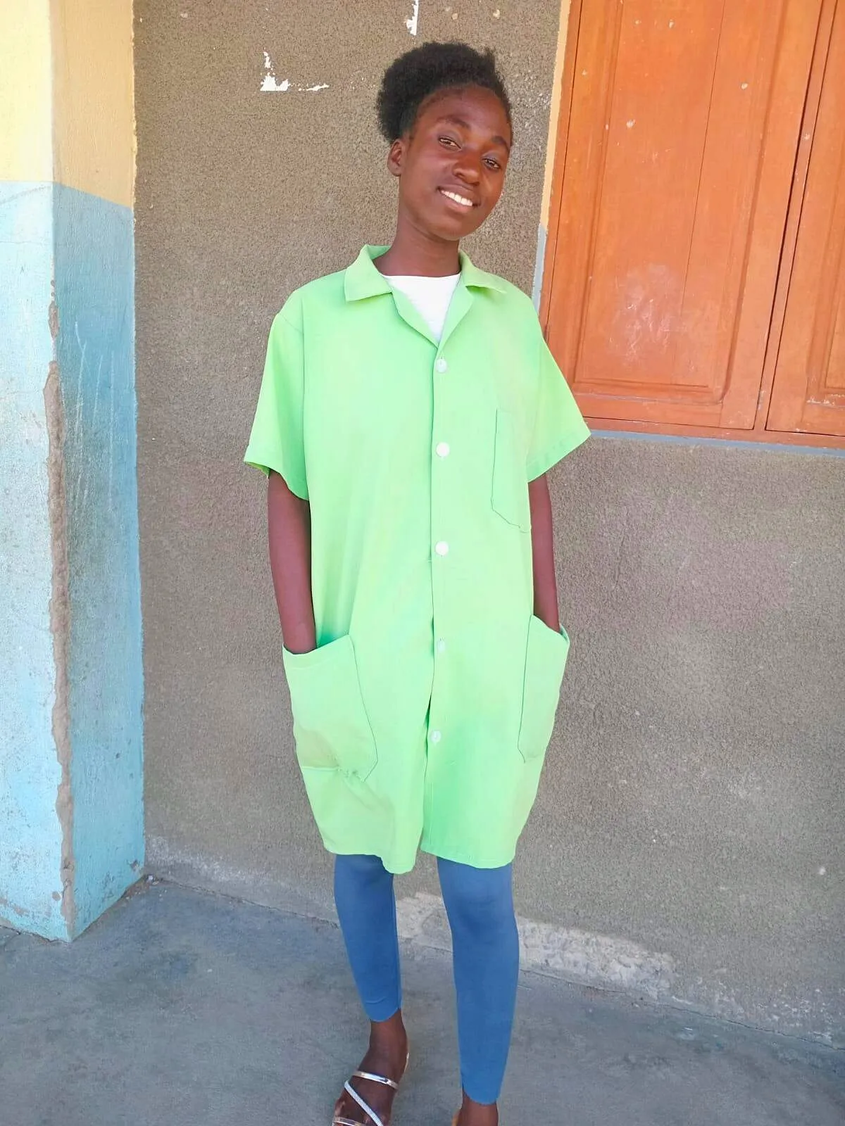 Florência Alexandre Valoi, sorridente, de bata escolar verde-lima numa varanda, com as mãos nos bolsos