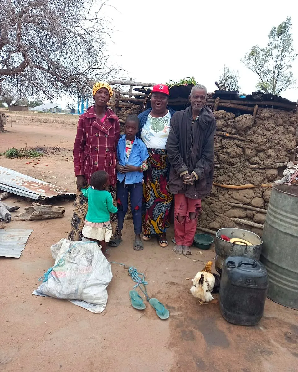 Uma família, com um menino, uma criança pequena, duas mulheres e um idoso, de pé diante de uma casa de pau e barro