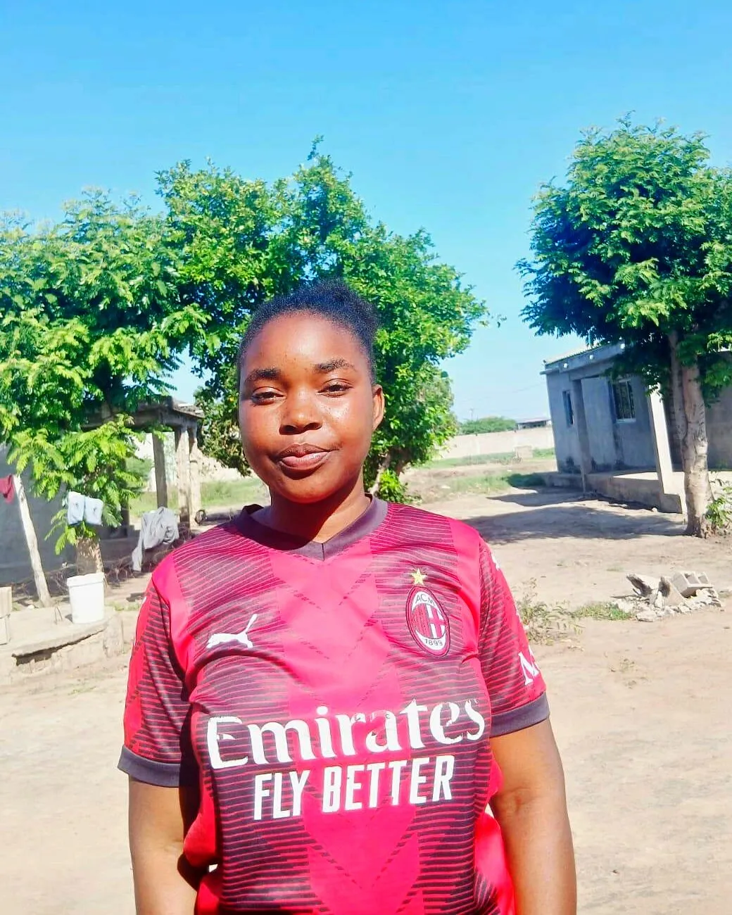 Misca Sérgio Ubisse, com um leve sorriso, de camisola de futebol vermelha e preta num quintal de areia entre duas árvores