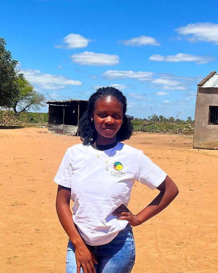 Samira Mandlate, sorridente, com a mão na anca, de T-shirt branca do Avança+ e calças de ganga, em terra vermelha diante de uma casa pequena