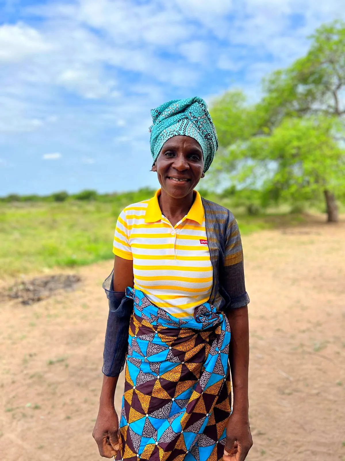 Uma mulher sorridente de lenço turquesa, polo às riscas e capulana estampada, num caminho de areia diante de uma grande árvore